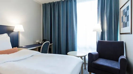 Bed with white sheets sits in a hotel room; a desk with a lamp and chair is nearby. Blue curtains cover the window, complemented by a blue armchair.