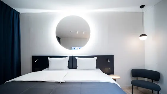 A modern bedroom featuring a large, neatly made bed with white and dark blue bedding, beneath a round, illuminated mirror. A small chair and hanging light fixture complement the minimalist decor.