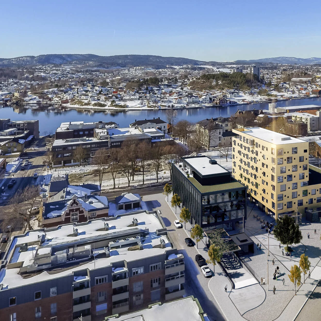 En klynge af moderne bygninger står nær en flod, omgivet af snedækkede gader og træer. I baggrunden strækker et malerisk bylandskab sig, med bakker synlige i det fjerne.