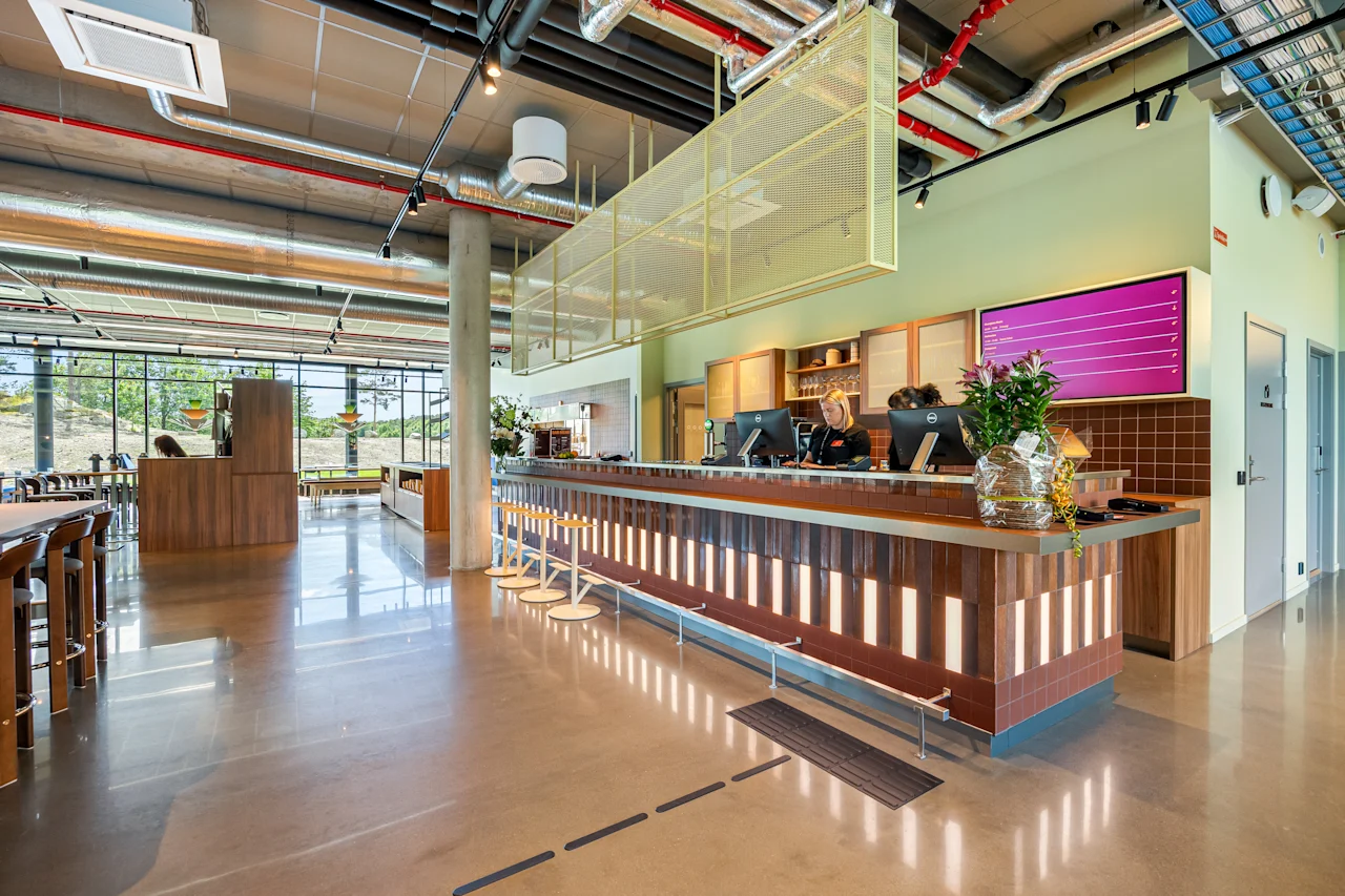 A modern and spacious hotel lobby with a long reception counter, bar stools, and large windows overlooking an outdoor area.
