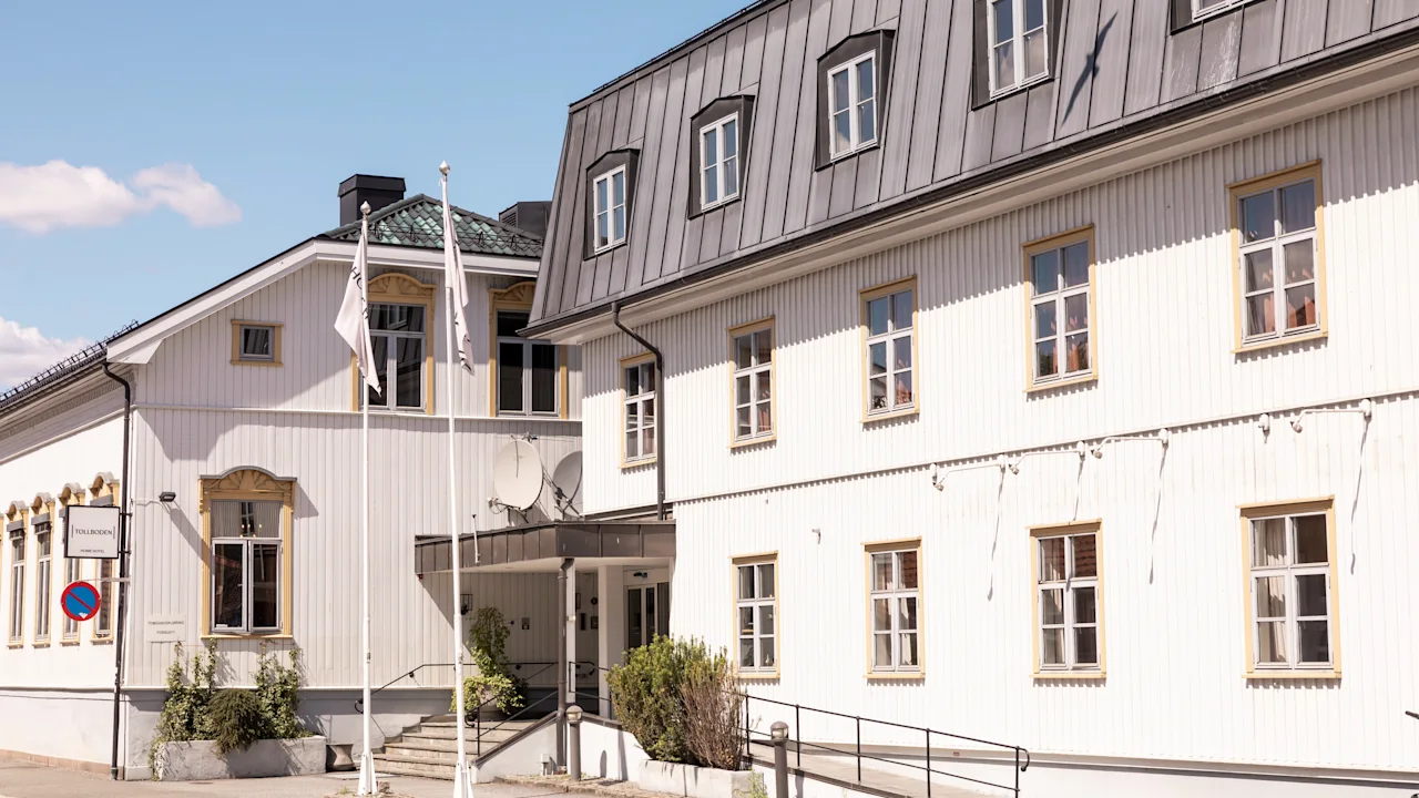 En hvid, toetagers træbygning med flere vinduer og metaltag befinder sig på en solrig gade med klar blå himmel. Ingen tekst er synlig.