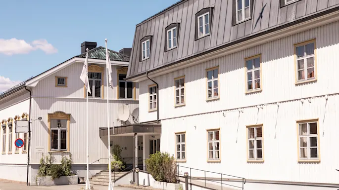 A white, two-story wooden building with multiple windows, metal roof, and a small entrance is situated on a sunny street with a clear blue sky. No text is visible.