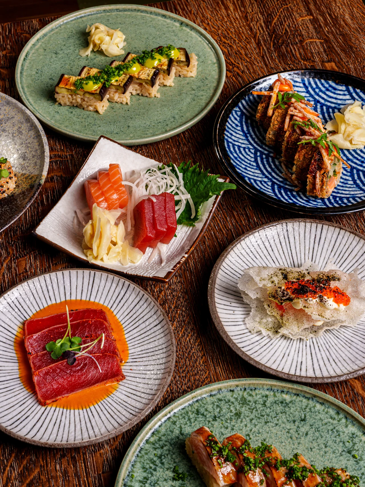 Sushi and sashimi dishes on various plates sit on a wooden table, featuring different types such as sushi rolls, sashimi with wasabi and ginger garnish.