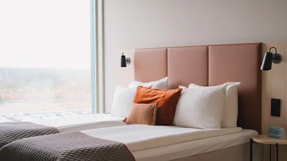 A neatly made twin bed with brown and white pillows rests against a padded headboard. Black wall-mounted lamps flank the bed, and a large window reveals a rainy landscape.