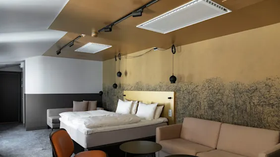 Et moderne hotellrom har en oppredd seng, to lenestoler, en sofa og et lite bord, mot en dekorativ vegg med abstrakte mønstre.