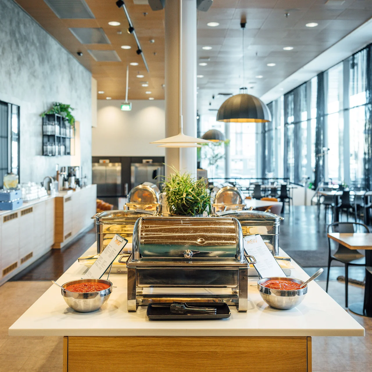 Buffet setup with chafing dishes on a wooden counter, flanked by bowls of sauce. Modern dining area with tables, chairs, and large windows in the background.