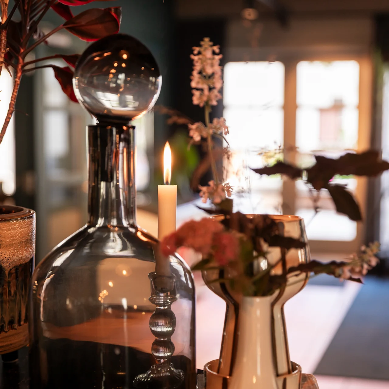 En glasflaske med en sfærisk prop står ved siden af et stearinlys på et bord. Det er omgivet af planter i et blødt oplyst rum med store vinduer i baggrunden.