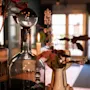 En glasflaske med en sfærisk prop står ved siden af et stearinlys på et bord. Det er omgivet af planter i et blødt oplyst rum med store vinduer i baggrunden.
