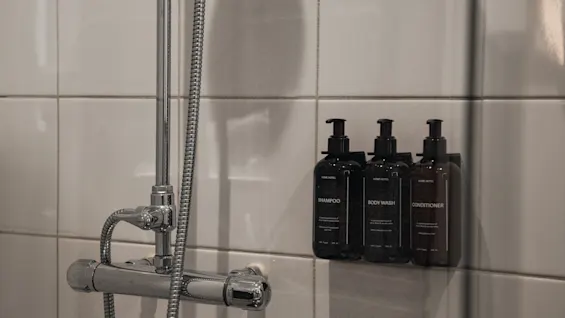 En dusch med vita kaklade väggar, en kromkran och tre väggmonterade flaskor märkta Schampo, Kroppstvål och Balsam.