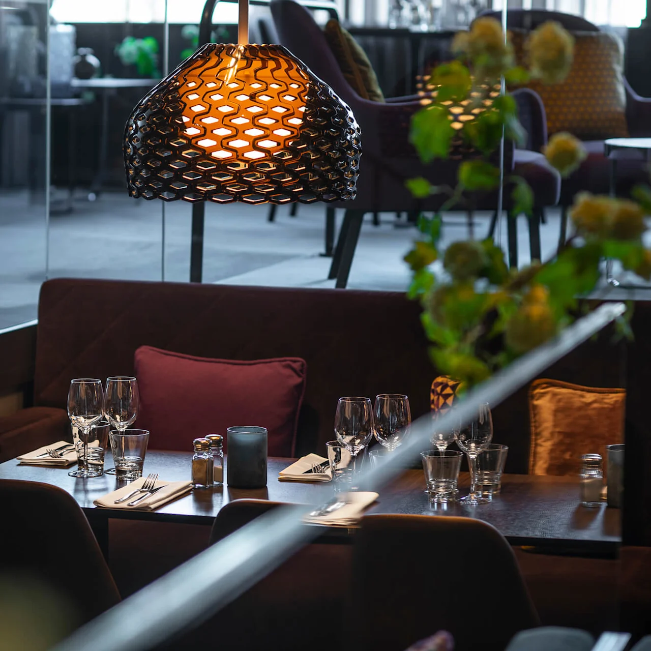 Matbord dukat med glas och bestick, under en dekorativ pendellampa, omgivet av plyschstolar i en modern restaurangmiljö.