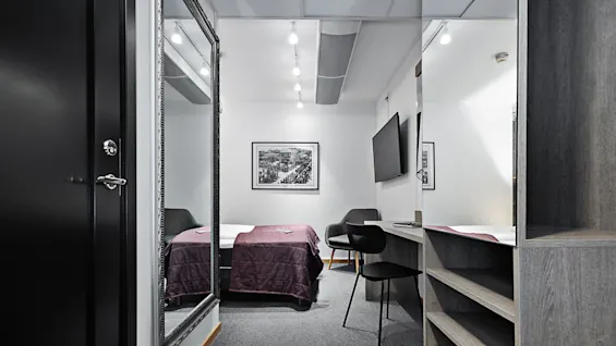 Ett litet hotellrum med en säng täckt av ett lila täcke. En stol, TV, stor spegel och väggkonst kompletterar den moderna inredningen.