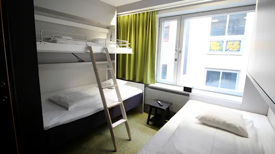 Valoisa hotellihuone, jossa on kerrossänky ja yhden hengen sänky suuren ikkunan ja vihreiden verhojen vieressä.
