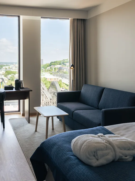 A bright and inviting hotel room featuring a cozy bed, a sofa, a work desk, and large windows with a scenic city view.
