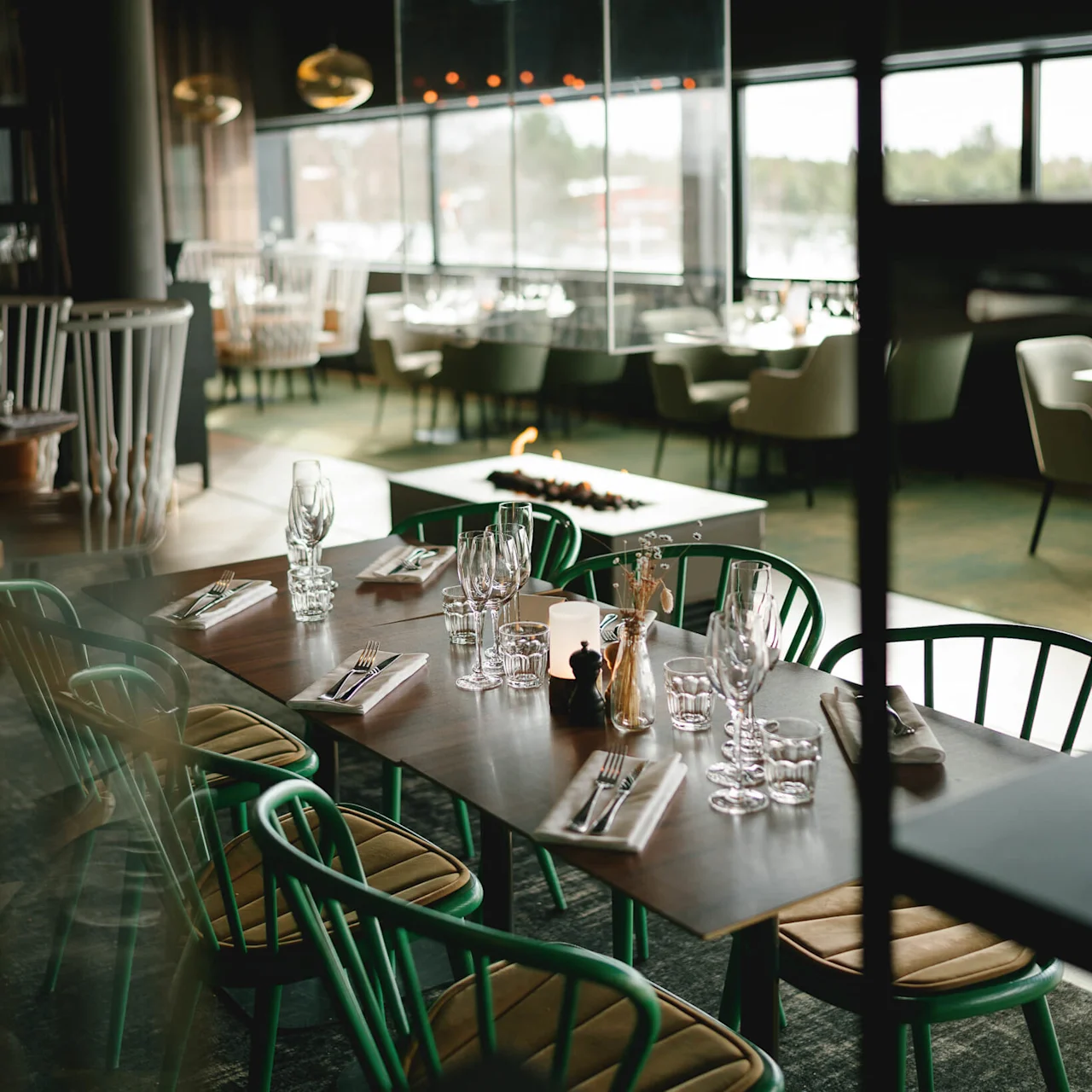 Ett bord med gröna stolar är dukat med glas, bestick och servetter i en modern restaurang. En glasomsluten eldstad och andra sittplatser skapar en mysig atmosfär.