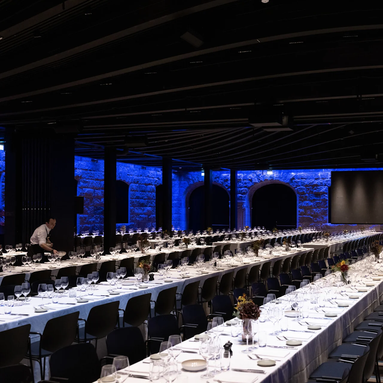 Long banquet tables set with glasses and plates line a dimly lit hall. A person arranges items on a table, with stone walls illuminated in blue light.  