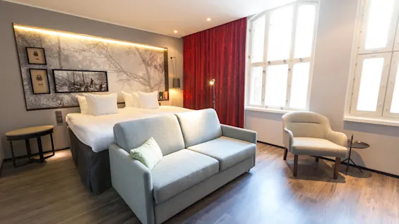 A modern hotel room with a bed, sofa, and art on the walls, illuminated by natural light from large windows.