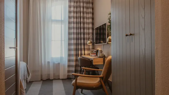 A cozy hotel room with a desk, chair, and TV, bathed in natural light from a large window with patterned curtains.