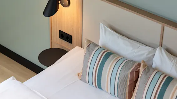 Ett mysigt hotellrum med en prydligt bäddad säng, vita lakan och färgglada randiga kuddar. En modern vägglampa lyser upp sänggaveln i trä.