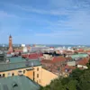 En klocktorn i röd tegelsten reser sig över stadens färgglada hustak, med blå himmel och avlägset hav i bakgrunden.