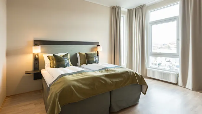 A neatly made bed with plush pillows in a bright, modern bedroom, large window providing a city view.