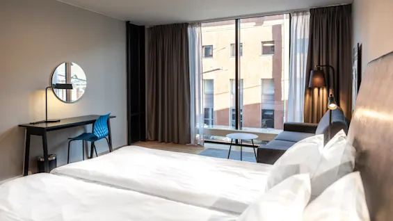 A modern hotel room features two neatly made beds, a desk with a blue chair, and a large window showcasing an urban building, creating a cozy, minimalist atmosphere.