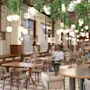 Restaurant interior features hanging green plants, round lamps suspended from the ceiling. Wooden tables are arranged with upholstered chairs. People are socializing. A sign reads "OPEN".