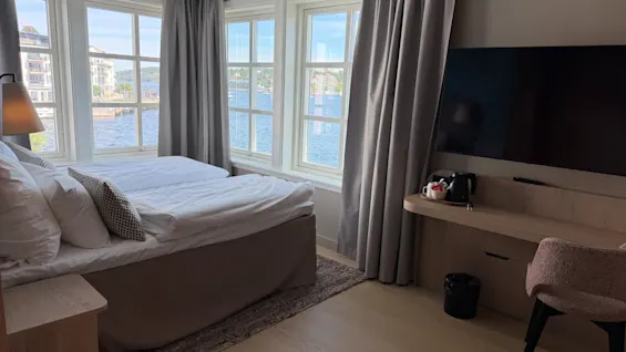 A cozy hotel room with a bed, large windows offering a scenic water view, a TV, and a small desk area.