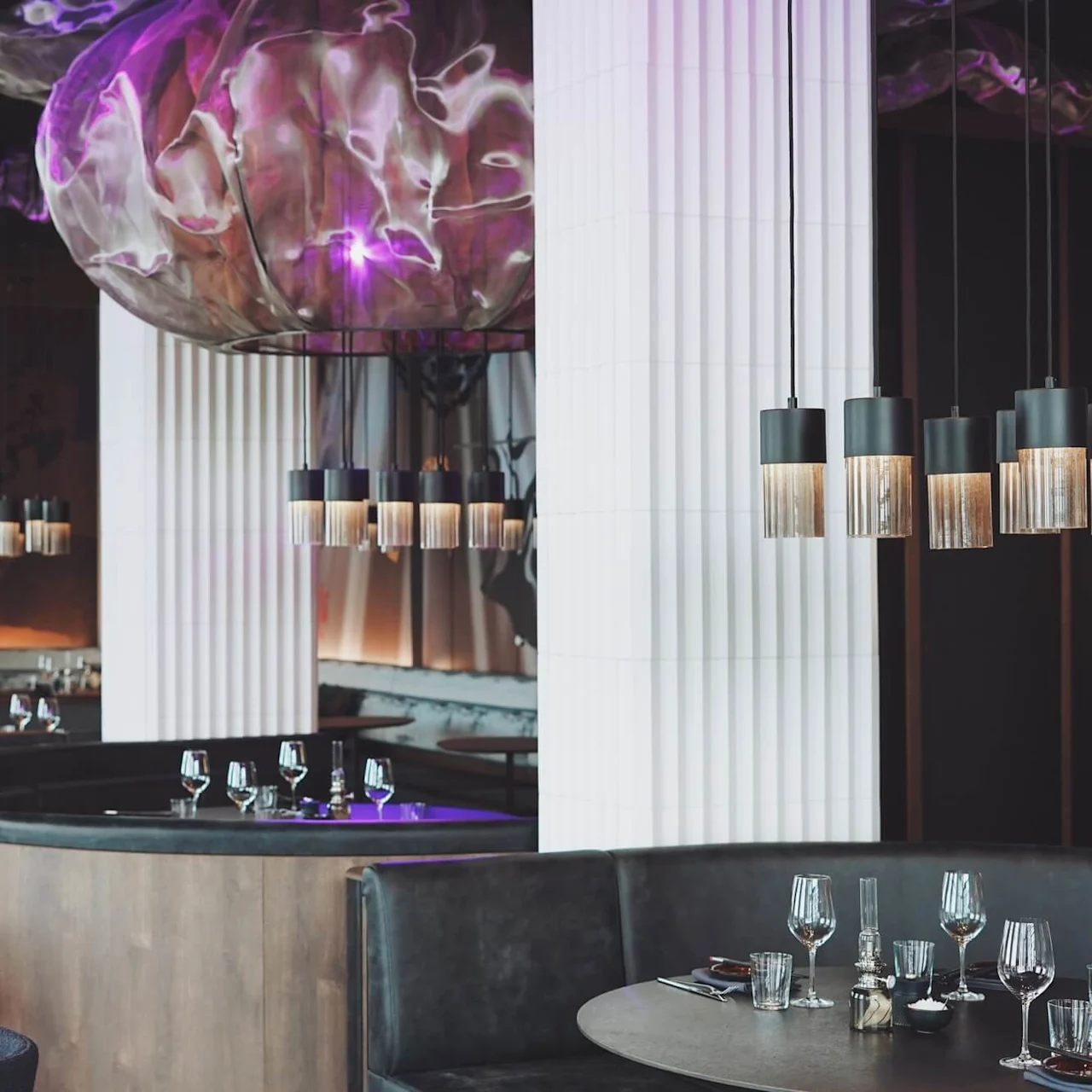 Elegant restaurantinteriør med hengende pendellamper som lyser opp runde bord dekket til middag. Omgitt av mørke sitteplasser og store hvite søyler. Lilla belysning fremhever den stilige atmosfæren.