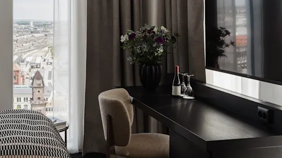 Et moderne hotellrom med skrivebord, stol, blomster og drikke, som tilbyr panoramautsikt over byen fra et stort vindu.