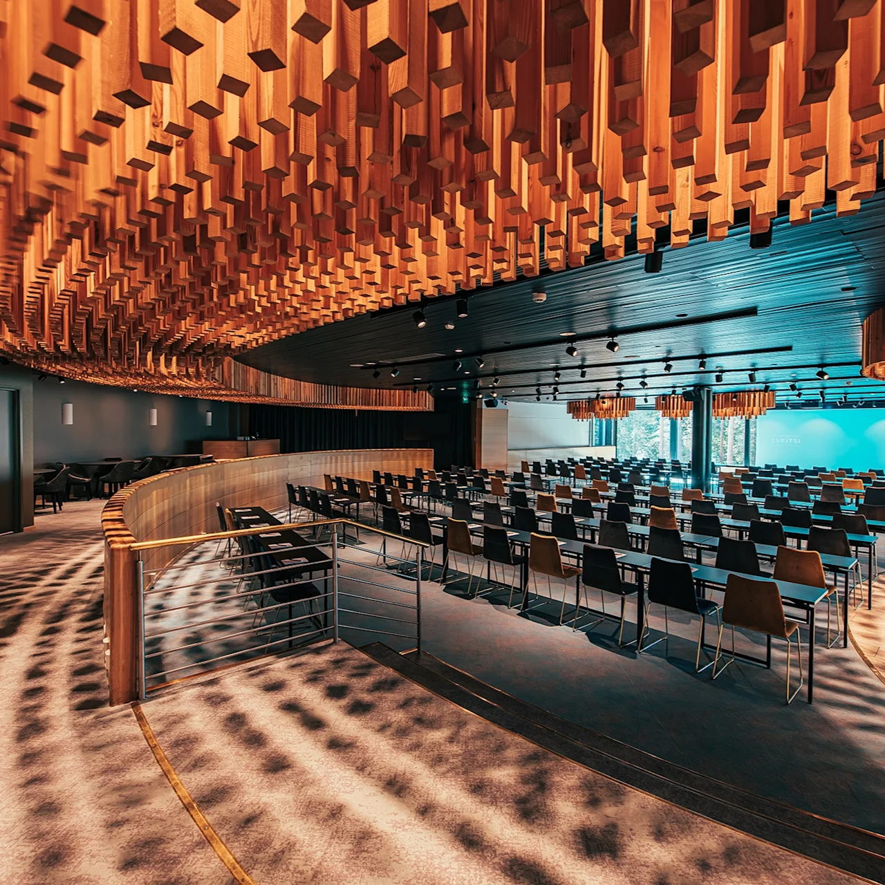 Rows of empty chairs face a stage in a modern auditorium. Overhead, a ceiling features suspended wooden panels. Soft lighting and shadows create a warm atmosphere in the spacious room.