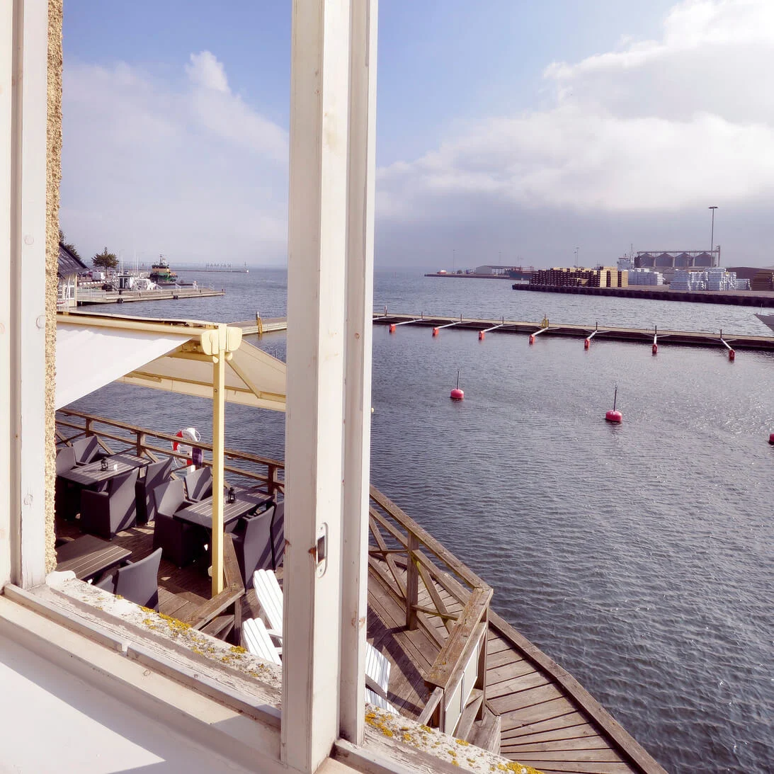Outdoor terrace by the water, with chairs and tables under an umbrella, viewed through an open window. A yacht and buoys are visible on the calm sea.