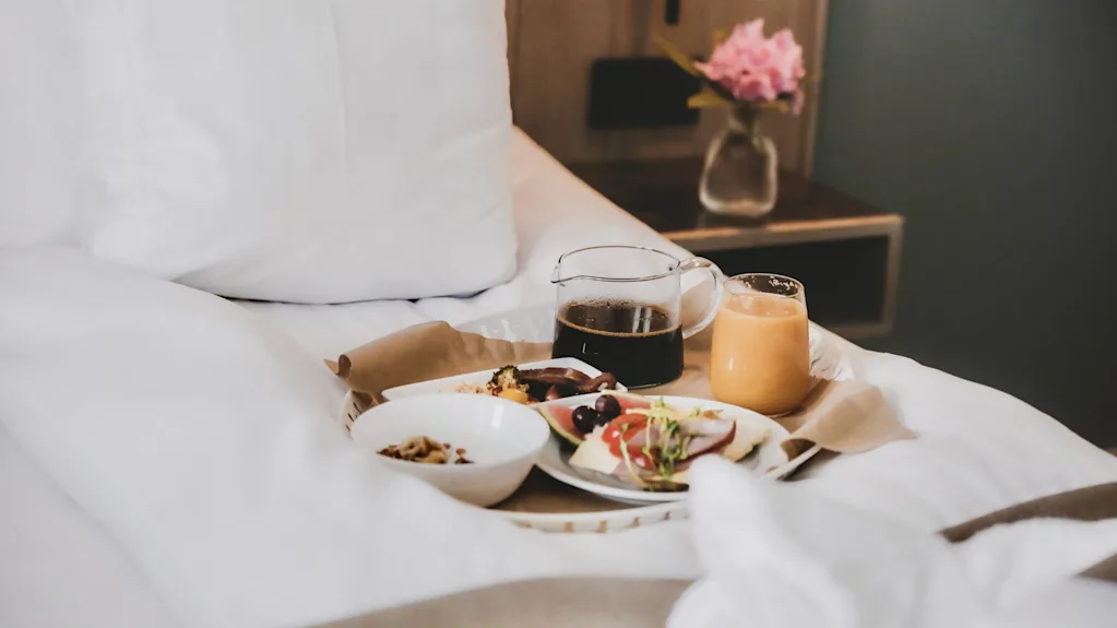 Breakfast tray on bed, featuring coffee, juice, and a variety of food items. A vase with pink flowers sits on a bedside table nearby.