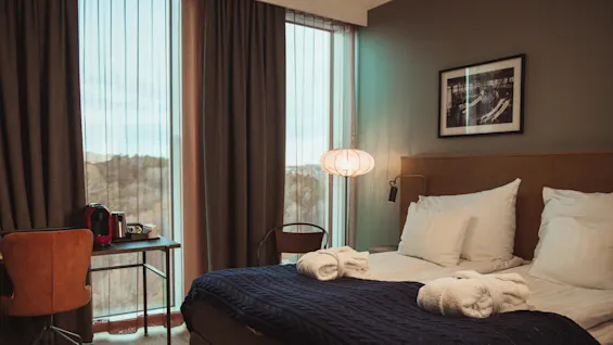 En prydligt bäddad hotellsäng med vita lakan och blå pläd har vikta handdukar. Ett närliggande skrivbord med stol och kaffebryggare står bredvid stora fönster med gardiner.