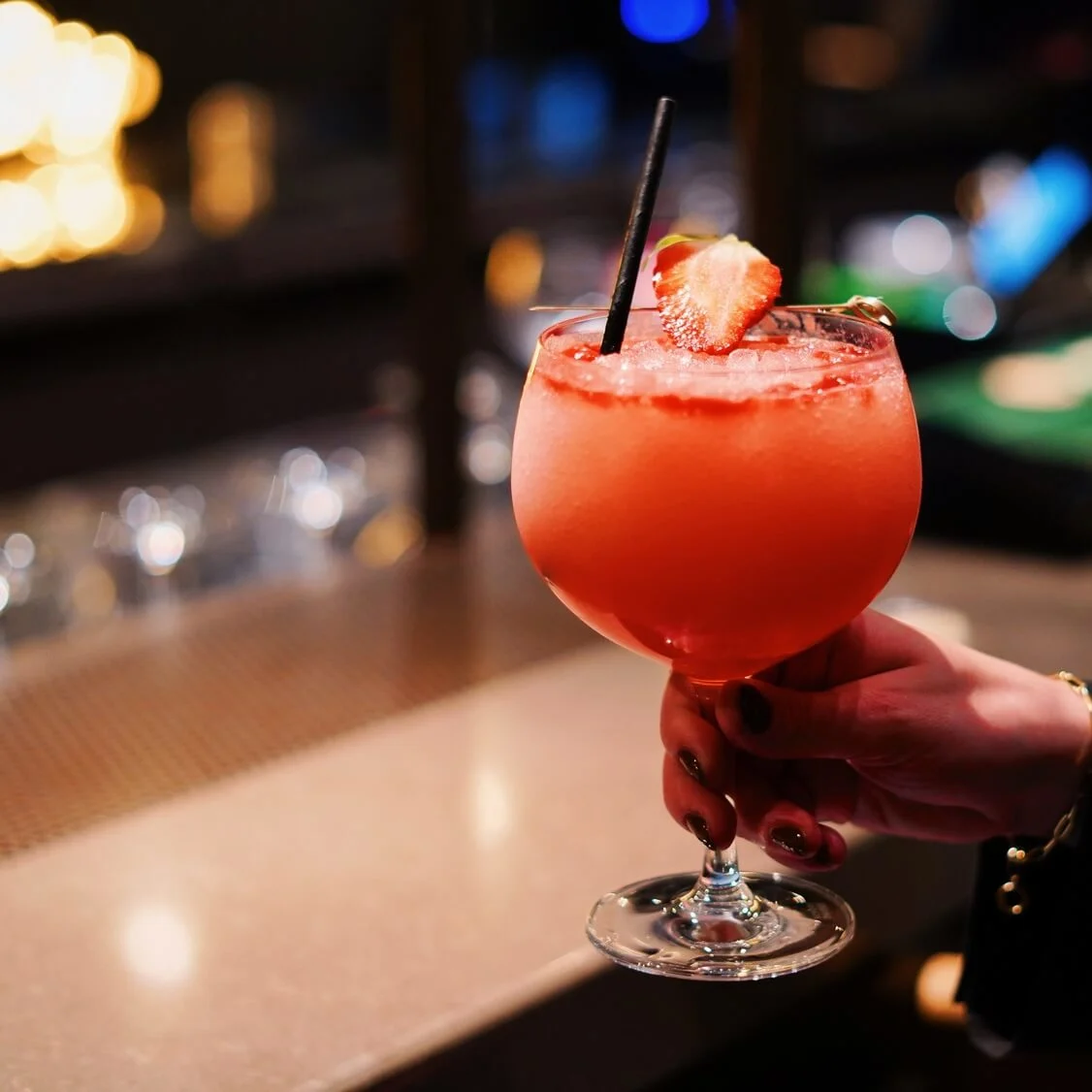 A strawberry cocktail garnished with a slice of strawberry is held by a hand over a bar counter, in a dimly lit bar setting.