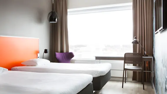 To pænt redte enkeltsenge står i et lyst hotelværelse med lyst trægulv. En lilla stol, skrivebord og lampe står nær et stort vindue med brune gardiner.