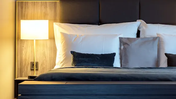 En seng med hvite og grå puter er opplyst av en moderne lampe på et sengebord av tre i et svakt opplyst soverom.