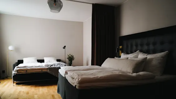 To pænt redte senge står i et svagt oplyst rum med trægulv, omgivet af enkle møbler, inklusive en lampe og en blomsterkrukke.