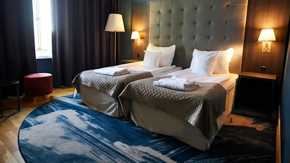 Two neatly made twin beds with white pillows and beige quilts stand in a hotel room, illuminated by warm bedside lamps, next to dark curtains and a patterned blue and beige rug.