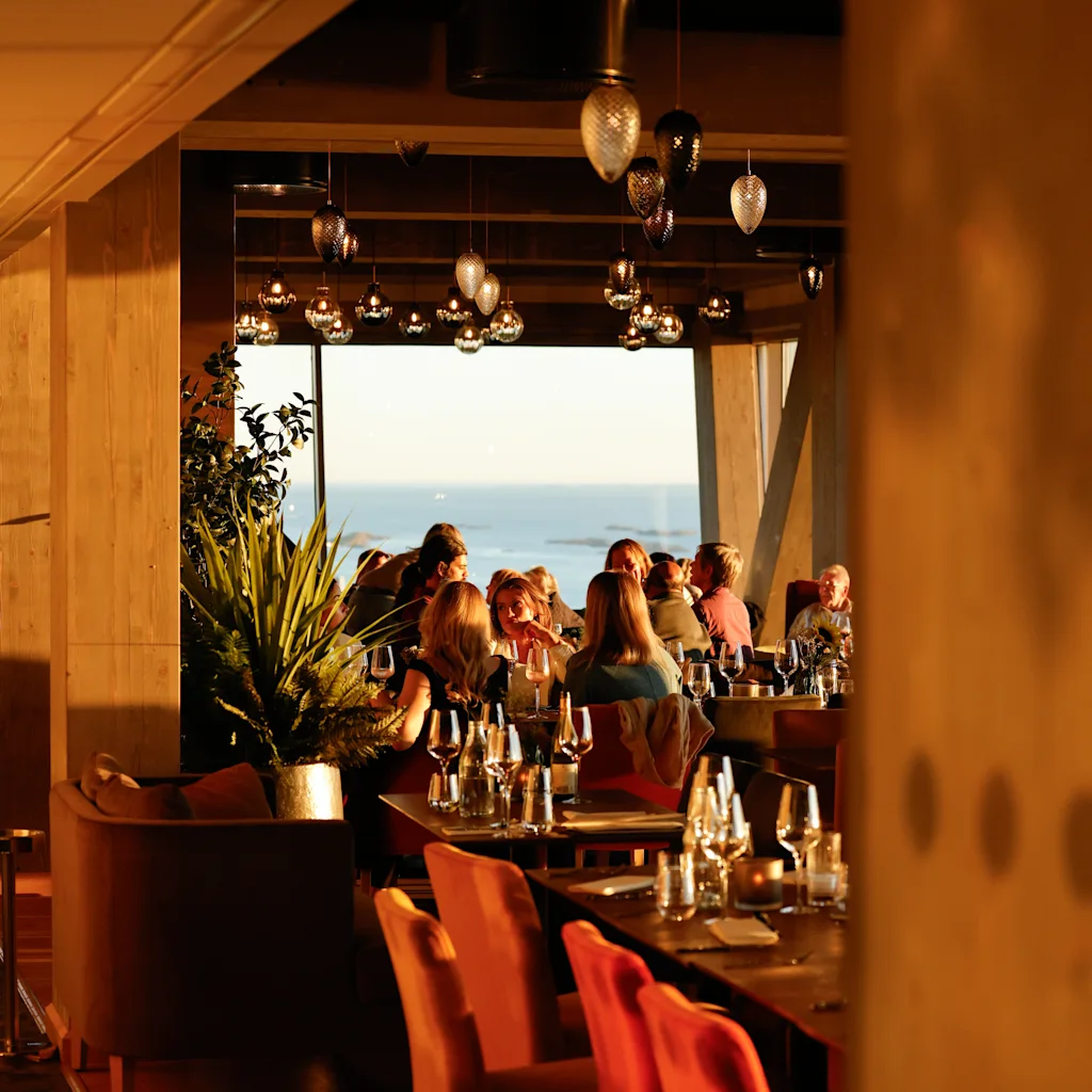 En gruppe mennesker spiser ved smakfullt dekkede bord i en varmt opplyst restaurant, med store vinduer som gir utsikt over havet, under et tak med hengende lys.