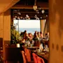 En gruppe mennesker spiser ved elegant dækkede borde i en varmt oplyst restaurant med store vinduer, der giver udsigt til havet, under et loft med hængende lamper.
