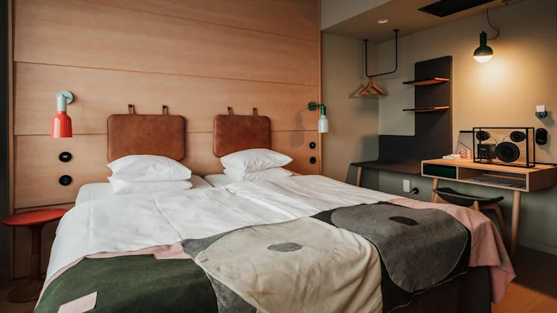 A neatly made double bed with white pillows and patterned blanket is in a modern hotel room. Wooden side table, red lamp, and desk area with lighting are nearby.