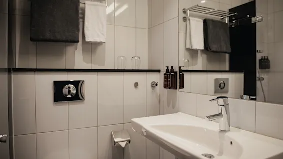 Ett rent och modernt hotellbadrum med vita kakelplattor, handfat, spegel, fräscha handdukar och toalettartiklar.