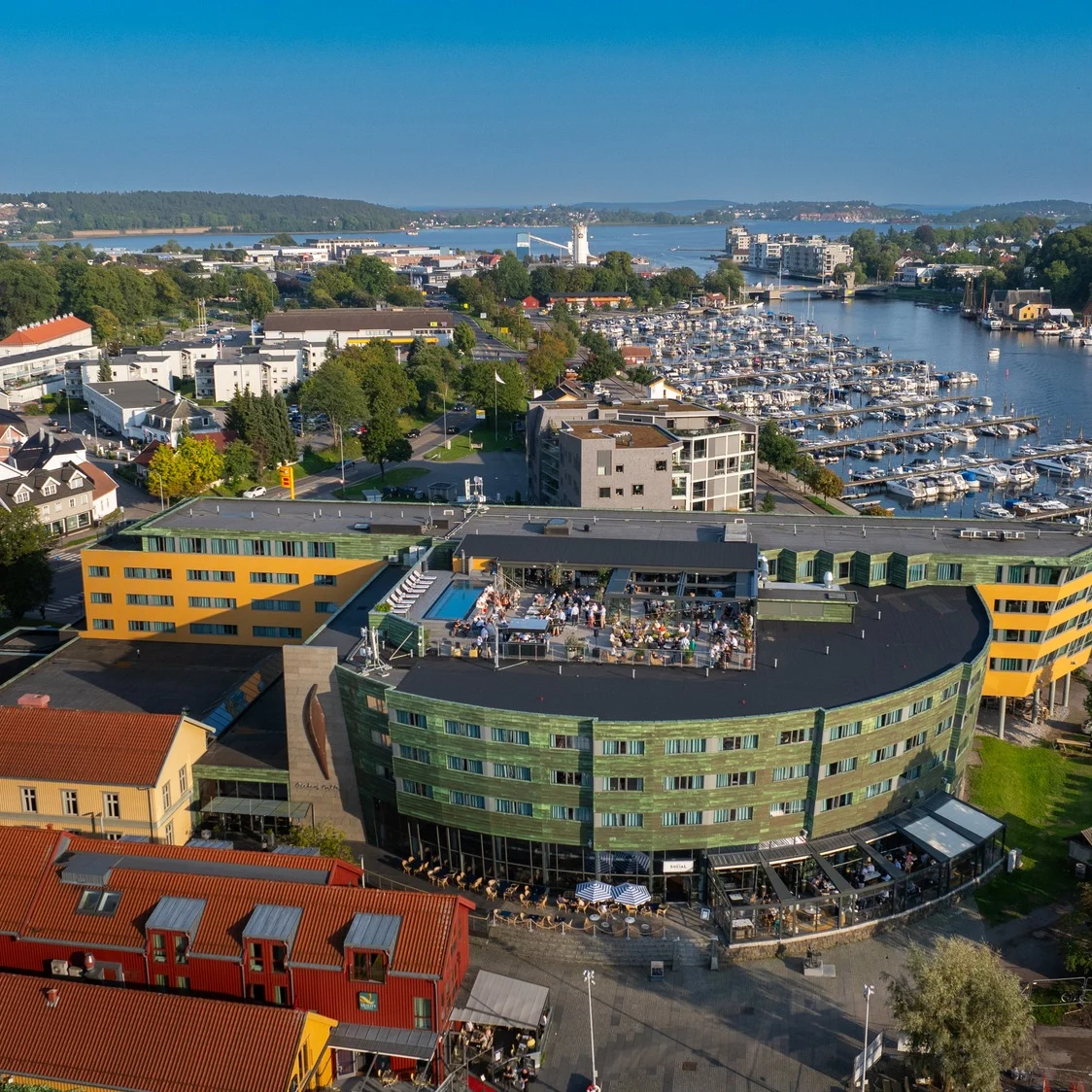 En grön och gul byggnad med takfester blickar ut över en marina full av båtar, omgiven av en kuststad med färgglada hus och frodig grönska under en klarblå himmel.