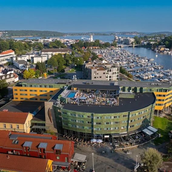 En grønn og gul bygning med taksamlinger skuer ut over en marina fylt med båter, omgitt av en kystby med fargerike hus og frodig grønt under en klar blå himmel.
