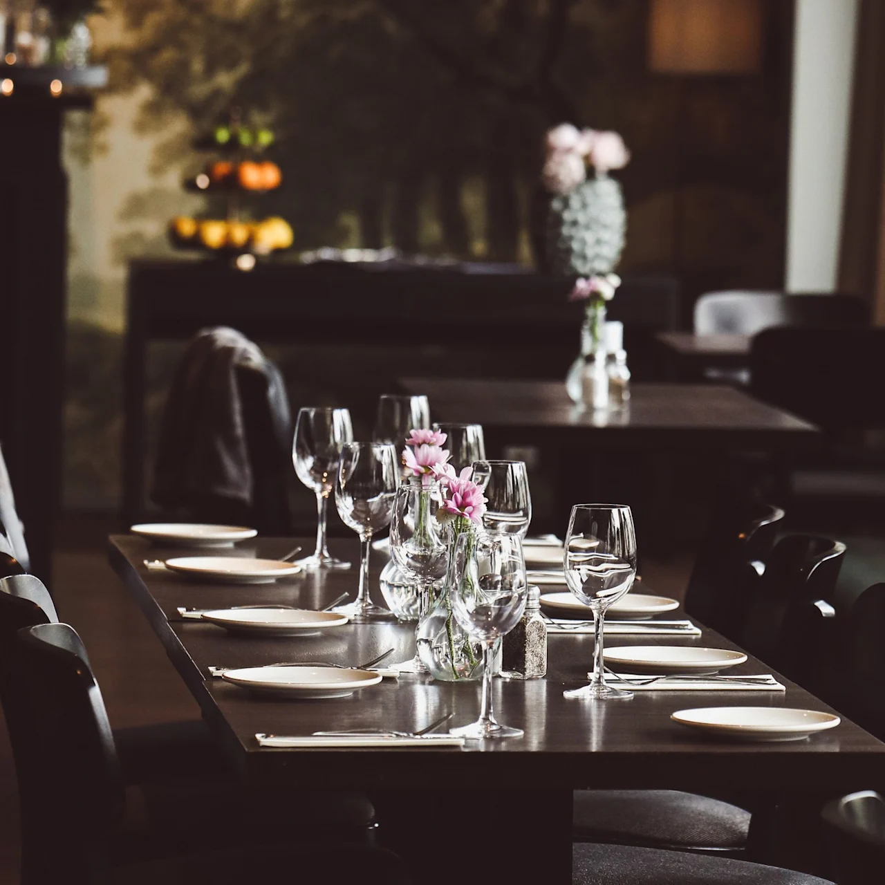 A dining table is elegantly set with plates, wine glasses, and pink flowers in vases; surrounded by dark chairs, in a dimly lit, sophisticated restaurant setting.