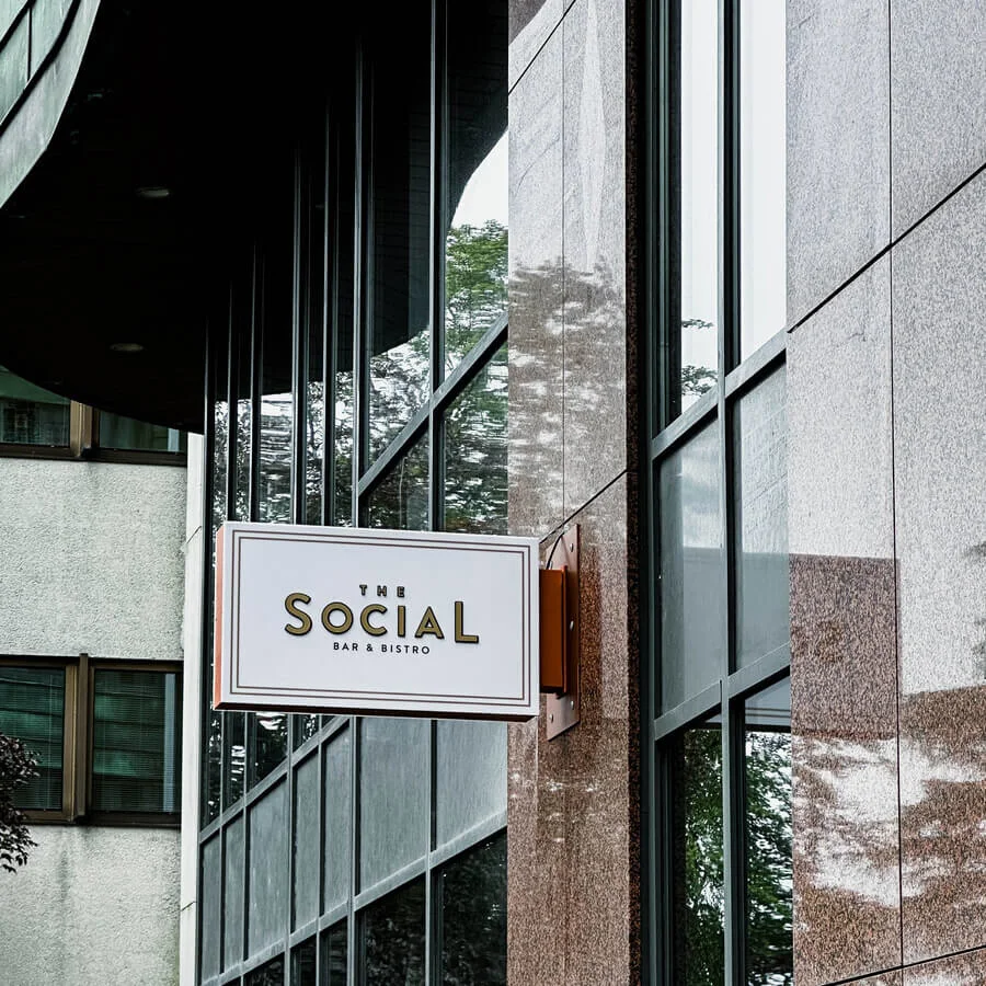 A rectangular sign shows "THE SOCIAL BAR & BISTRO" in gold letters, attached to a building with reflective glass and textured stone panels, surrounded by other urban structures.