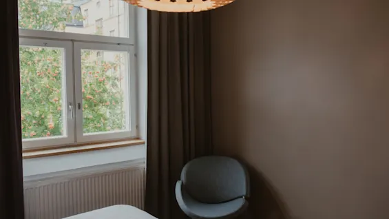 A cozy room at Home Hotel Uppsala with a window overlooking green trees, a bed, a modern chair, and a decorative ceiling light.