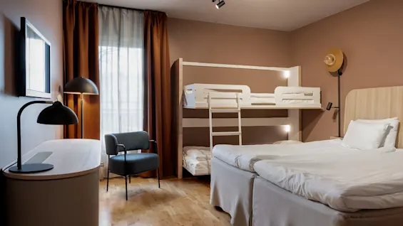 Ett mysigt hotellrum med en våningssäng, två enkelsängar, ett skrivbord med lampa och en väggmonterad TV, mot varma bruna väggar och trägolv.