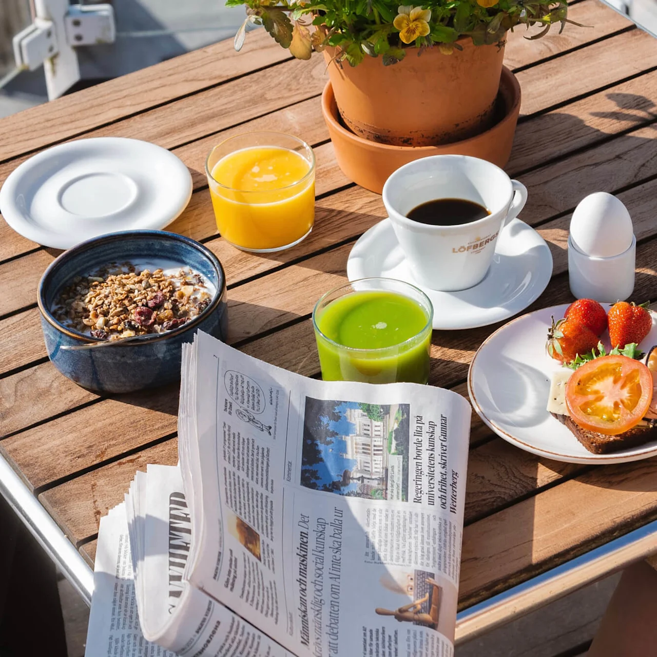 Et trebord har en frokostoppsett med kaffe, juice, en bolle med granola, og en avis. En potteplante legger til grønt. Tekst på kopp: "Löfbergs."