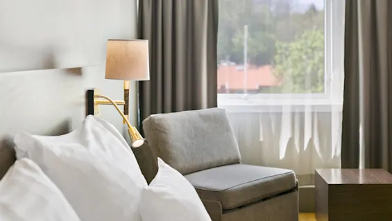 Viihtyisä hotellihuoneen nurkkaus, jossa sänky, yöpöytälamppu, nojatuoli ja ikkuna, josta näkyy vehreyttä.